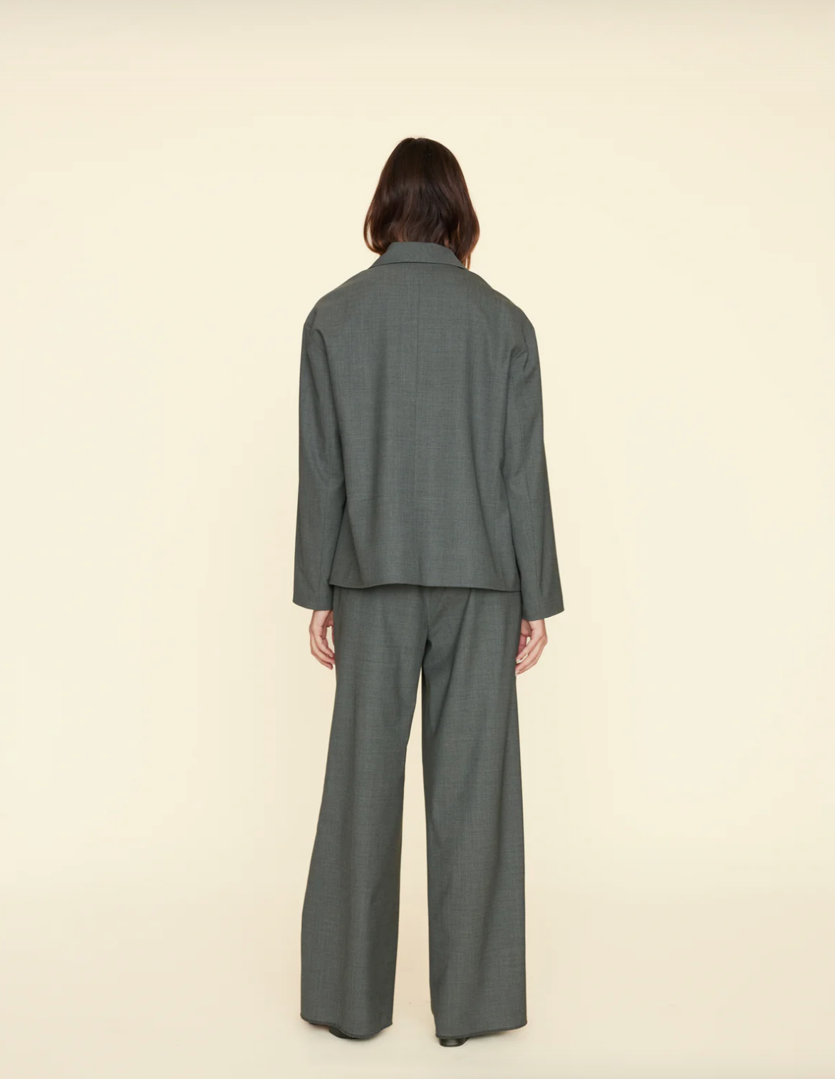 A person with shoulder-length hair is seen from behind, wearing a loose-fitting, dark gray pantsuit. The suit jacket is slightly oversized, offering a soft-tailored look, and the wide-legged Portlyn Pant by Xirena reaches the floor. Made from a wool blend, the ensemble stands out against the plain, light beige background.