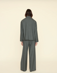 A person with shoulder-length hair is seen from behind, wearing a loose-fitting, dark gray pantsuit. The suit jacket is slightly oversized, offering a soft-tailored look, and the wide-legged Portlyn Pant by Xirena reaches the floor. Made from a wool blend, the ensemble stands out against the plain, light beige background.