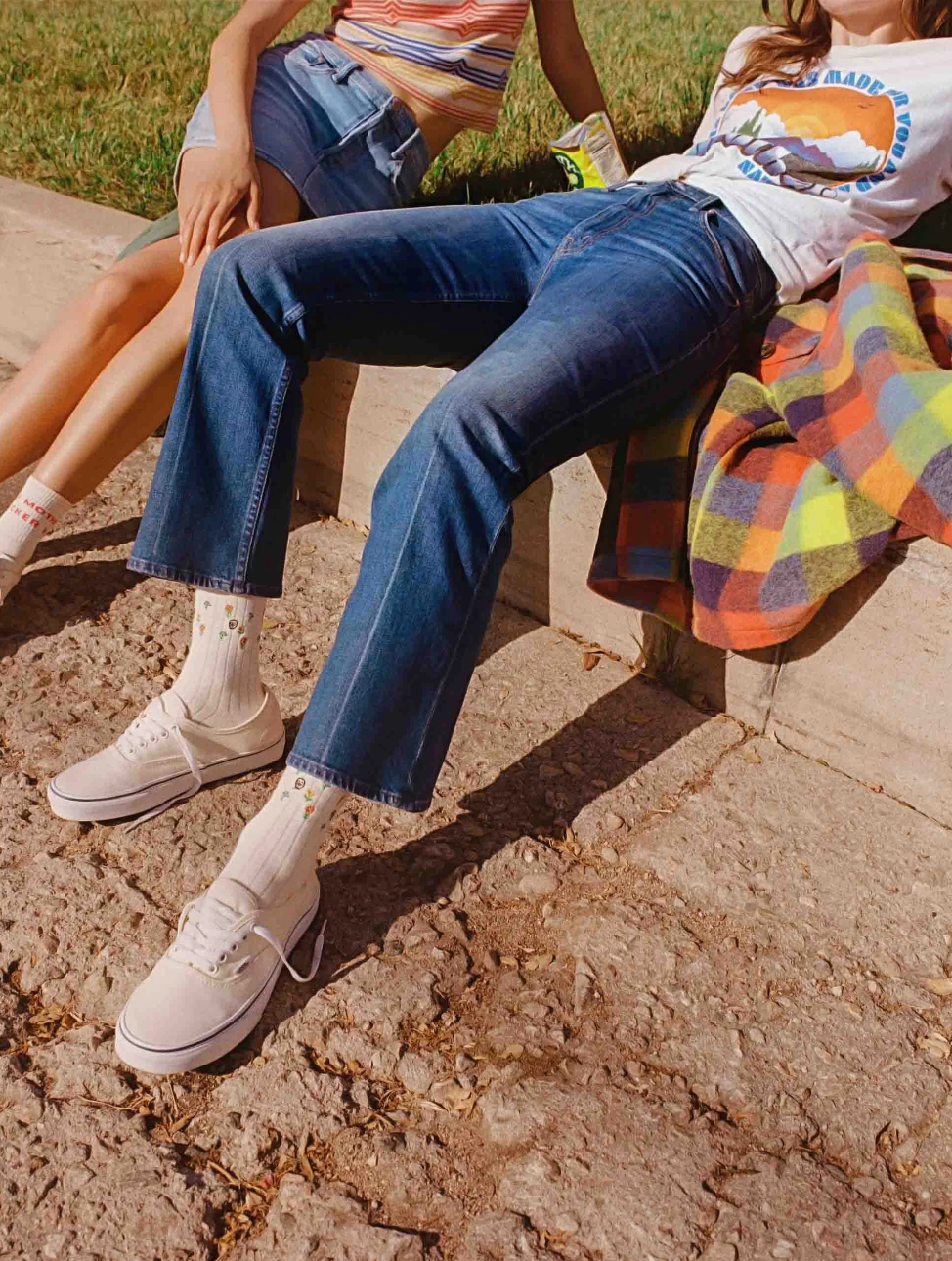 Two people sitting casually on a concrete ledge in a sunny outdoor setting. One wears a colorful striped tank top and denim shorts; the other sports classic blue wash Super High-Rise Jeans "The Scooter Ankle" by Mother, paired with a graphic tee and white sneakers. A vintage look blanket is spread next to them.