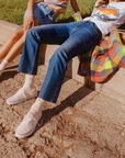 Two people sitting casually on a concrete ledge in a sunny outdoor setting. One wears a colorful striped tank top and denim shorts; the other sports classic blue wash Super High-Rise Jeans "The Scooter Ankle" by Mother, paired with a graphic tee and white sneakers. A vintage look blanket is spread next to them.