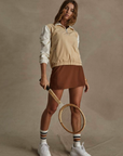 A woman stands against a plain backdrop wearing the MANSI DAWSON RUN JACKET by The Upside, complemented by a brown skirt and white sneakers with striped socks. She confidently holds a wooden tennis racket and gazes at the camera, embodying both style and function much like our moisture-wicking apparel.