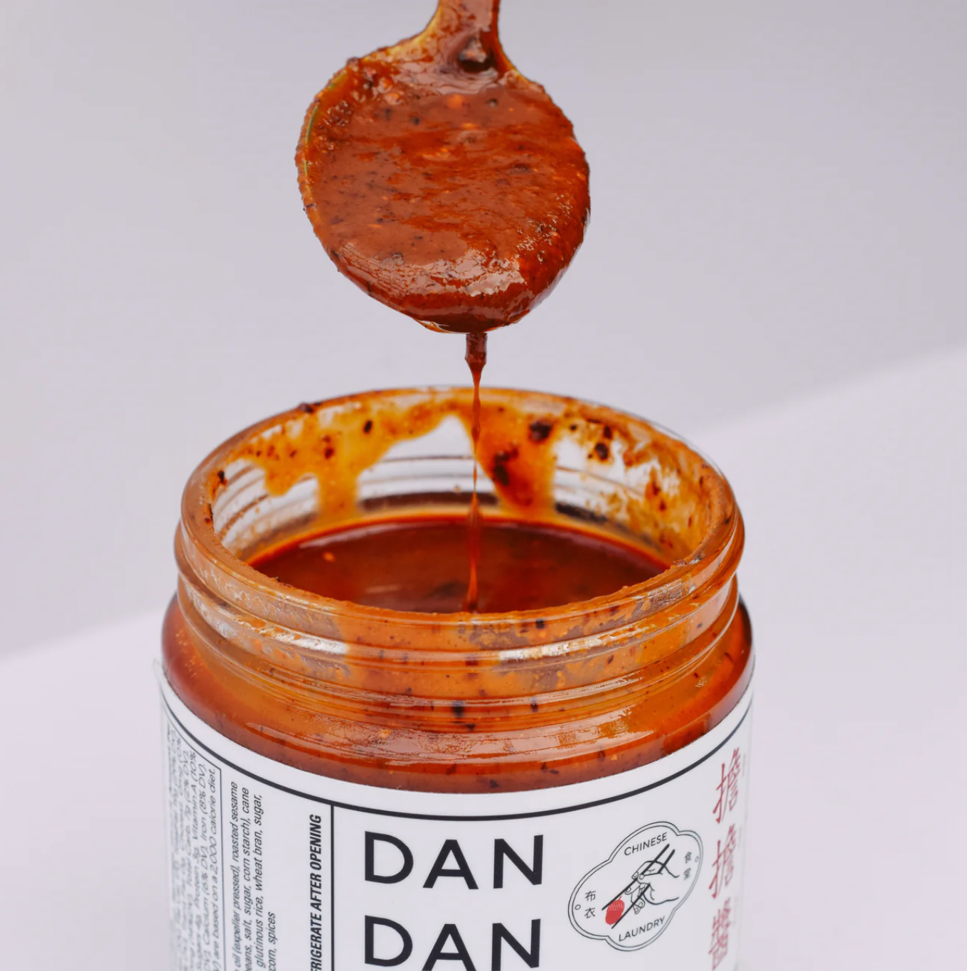 A spoonful of thick, red Dan Dan sauce being lifted from an open jar labeled "Faire Sichuan Spicy Dan Dan Sauce" with Chinese characters. The sauce, originating from Sichuan province, has a rich, textured appearance with visible spices and an irresistible umami flavor that enhances any dish.