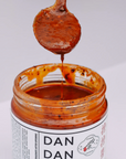 A spoonful of thick, red Dan Dan sauce being lifted from an open jar labeled "Faire Sichuan Spicy Dan Dan Sauce" with Chinese characters. The sauce, originating from Sichuan province, has a rich, textured appearance with visible spices and an irresistible umami flavor that enhances any dish.