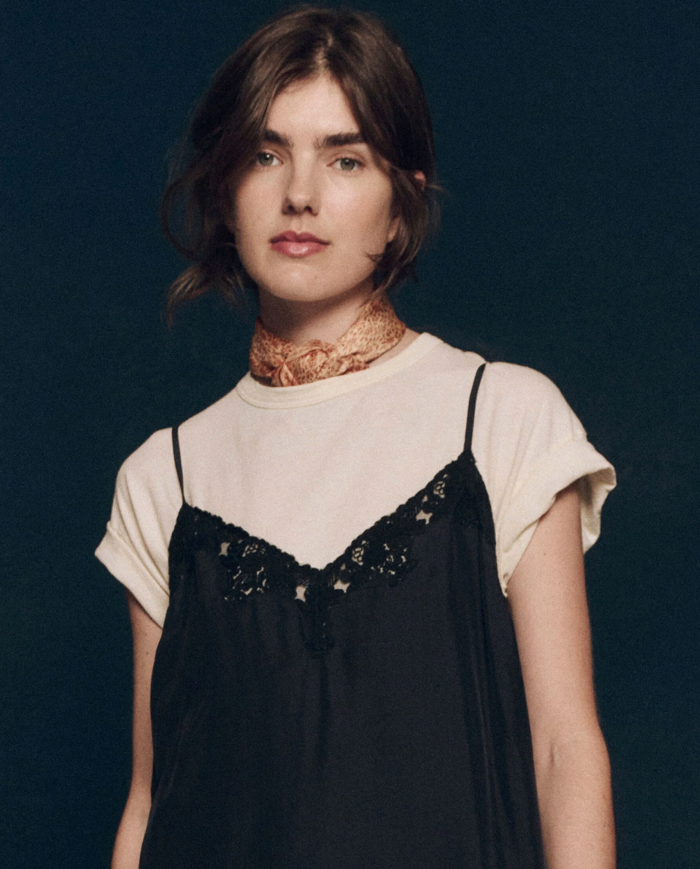 A person with shoulder-length dark hair is wearing a plain white T-shirt under The Legacy Dress by The Great Inc., featuring black lace trim and crafted from 100% silk, paired with a beige patterned scarf around their neck. They are standing against a dark background and looking towards the camera.