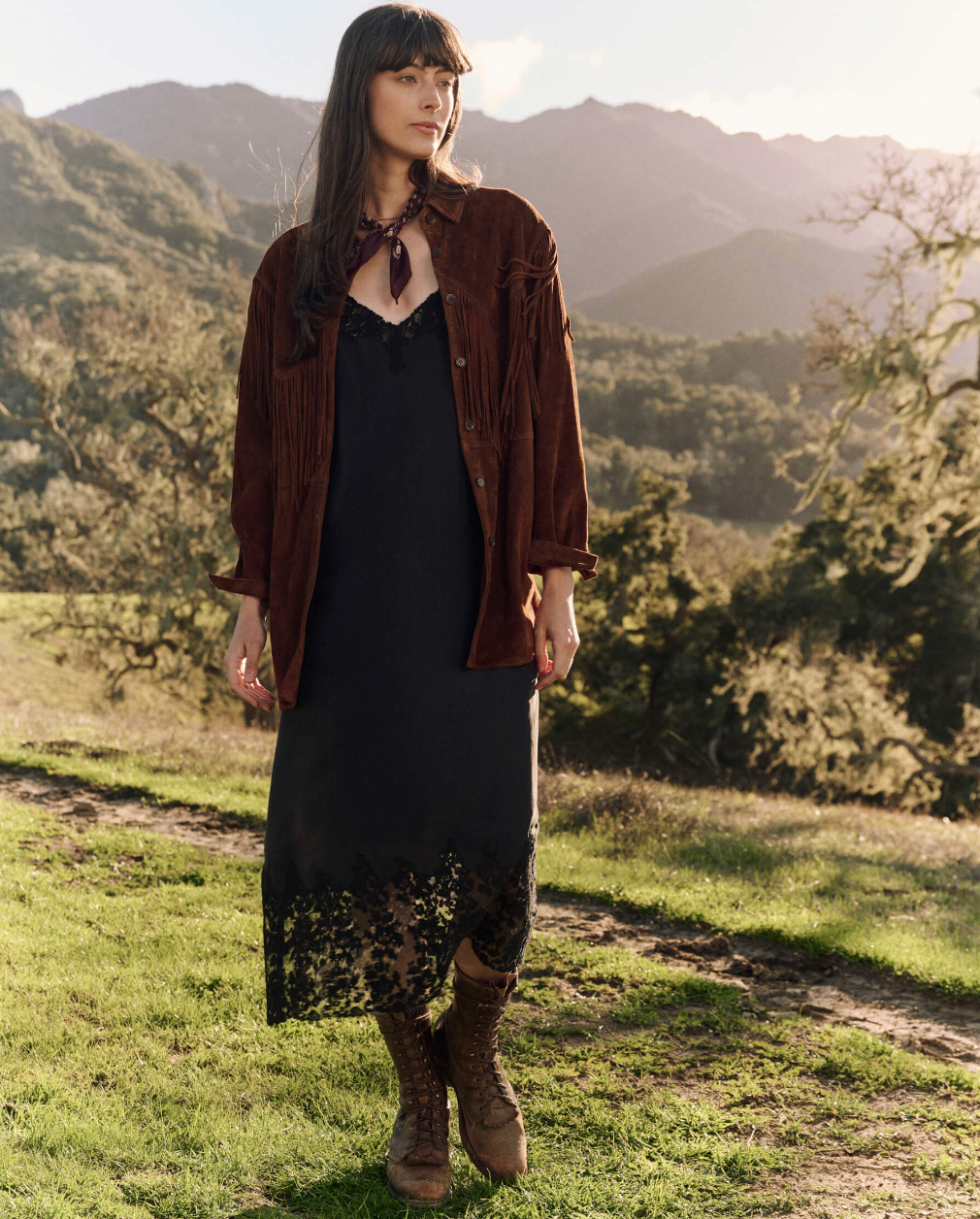 A woman stands outdoors in a rural setting with mountains in the background. She is dressed in a brown fringe jacket over "The Legacy Dress" by The Great Inc., featuring black lace trim and embroidered mesh lace. She accessorizes with a purple scarf around her neck and brown lace-up boots. The sun is shining, casting a warm light over her timeless fall look.