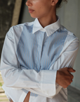 A person with short hair stands with arms crossed, wearing The BIB Shirt by Cissa, made of Egyptian Cotton Contrast, featuring a white dress shirt with vertical blue stripes on the front and a contrast bib. The person looks to the side with a neutral expression. The background includes a blurred structure and natural light.