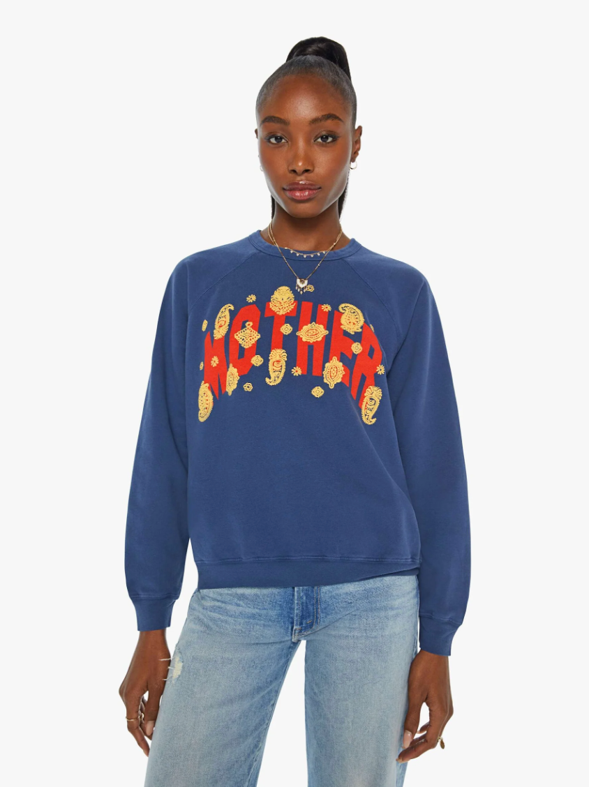 A person stands wearing The Biggie Concert raglan sweatshirt by Mother, crafted from super-soft cotton and showcasing a vibrant, 80s-inspired graphic in the center. Their hair is styled in a high bun, and they pair the sweatshirt with light blue jeans and delicate jewelry. The plain white background highlights the full focus on them and their outfit.