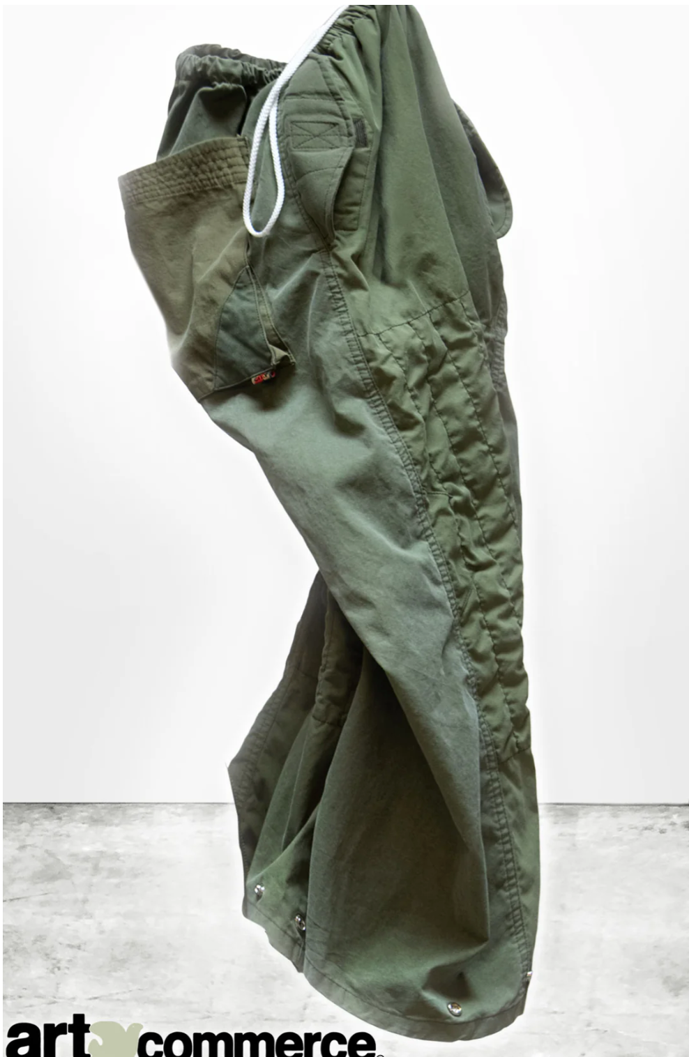 The RTU flap/snap KARATE BAG pant, designed by Free City (sparrow, LLC), hangs in mid-air against a light grey background. This pair of green karate pants is handmade in Los Angeles from vintage army tents and showcases an elastic waistband with a white drawstring, large side pockets, visible stitching, and buttoned cuffs at the ankles. The logo "art commerce" is partially visible at the bottom.