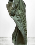 The RTU flap/snap KARATE BAG pant, designed by Free City (sparrow, LLC), hangs in mid-air against a light grey background. This pair of green karate pants is handmade in Los Angeles from vintage army tents and showcases an elastic waistband with a white drawstring, large side pockets, visible stitching, and buttoned cuffs at the ankles. The logo "art commerce" is partially visible at the bottom.