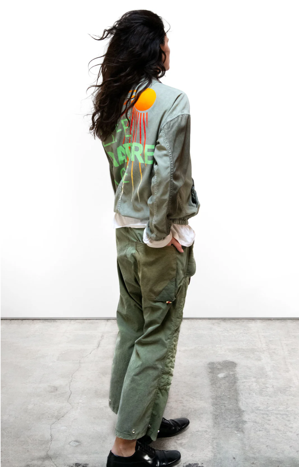 A person with long, wavy hair stands on a concrete floor, facing away from the camera. They wear an olive green jacket made from vintage army tents adorned with colorful abstract designs and matching Free City RTU flap/snap KARATE BAG pants. The person's stance is relaxed, and they are also wearing black shoes.