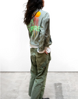 A person with long, wavy hair stands on a concrete floor, facing away from the camera. They wear an olive green jacket made from vintage army tents adorned with colorful abstract designs and matching Free City RTU flap/snap KARATE BAG pants. The person's stance is relaxed, and they are also wearing black shoes.