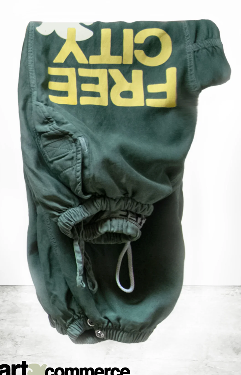 A pair of Flap/Snap LG MATTE/SATIN AirJump Pants from Free City (sparrow, LLC) in a custom sunfade wash green is hanging upside down on a white rectangular platform. The unisex sweatpants, featuring "FREE CITY" in large yellow lettering and an elastic waist and cuffs, are set against a simple grey and white gradient background. The text "art commerce" partially appears at the bottom.