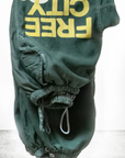 A pair of Flap/Snap LG MATTE/SATIN AirJump Pants from Free City (sparrow, LLC) in a custom sunfade wash green is hanging upside down on a white rectangular platform. The unisex sweatpants, featuring "FREE CITY" in large yellow lettering and an elastic waist and cuffs, are set against a simple grey and white gradient background. The text "art commerce" partially appears at the bottom.