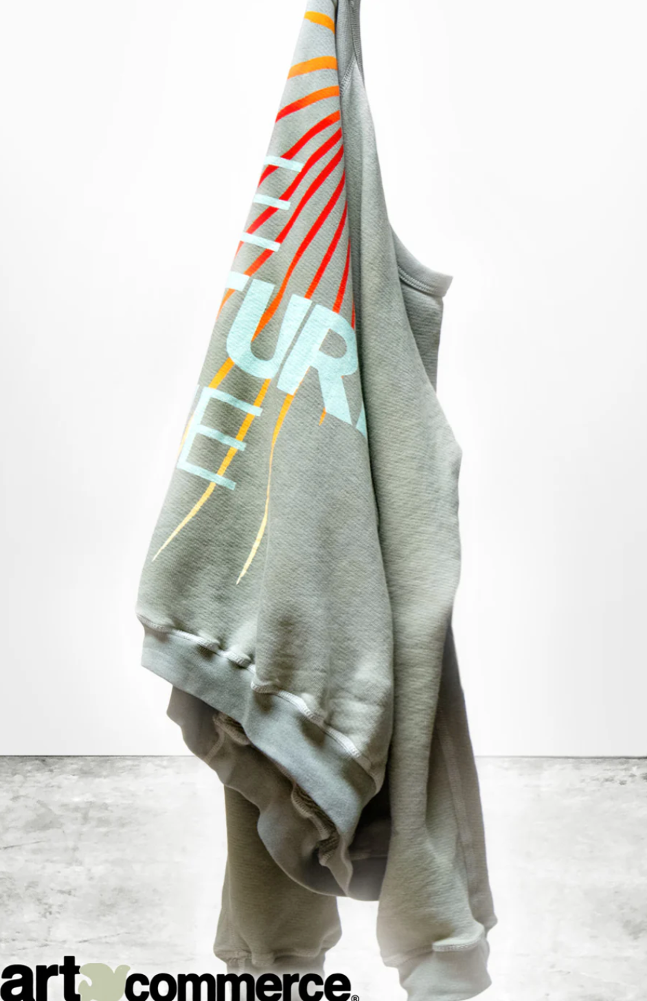 The LNLSUN SprYum Biggie Raglan hangs on a white background, presenting a lightweight gray French terry hoodie. This oversized unisex sweatshirt from Free City (sparrow, LLC) boasts a colorful, abstract design on the back with partially visible text &quot;FUTURE&quot; and &quot;COME&quot; in white and yellow. The concrete floor below adds to its appeal, while the &quot;art commerce&quot; logo is displayed in the bottom left corner.