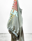 The LNLSUN SprYum Biggie Raglan hangs on a white background, presenting a lightweight gray French terry hoodie. This oversized unisex sweatshirt from Free City (sparrow, LLC) boasts a colorful, abstract design on the back with partially visible text "FUTURE" and "COME" in white and yellow. The concrete floor below adds to its appeal, while the "art commerce" logo is displayed in the bottom left corner.