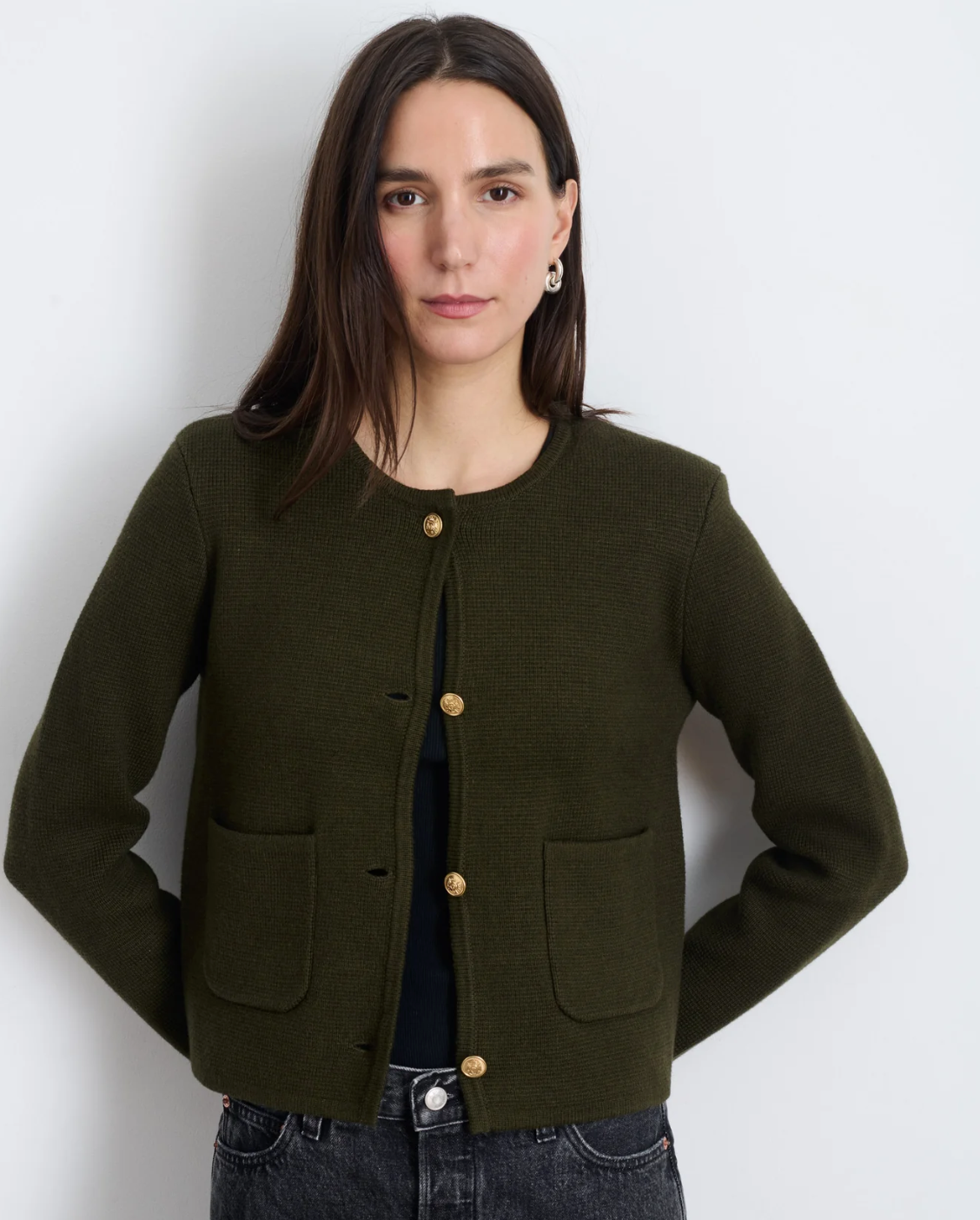 A person with long brown hair wearing the Paris Sweater Jacket by Alex Mill, which features brass crest buttons and front pockets, stands against a plain white background. They have a neutral expression and are dressed in dark jeans.