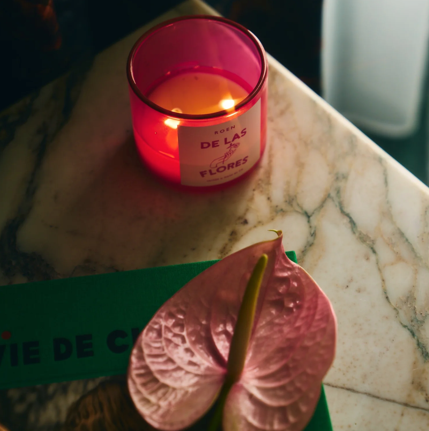 A lit ROEN Mexico City Candle by Faire in a glass holder sits on a marble surface next to a heart-shaped pink flower and a partially visible green book with the text "VIE DE C." The candle's label reads "ROSA DE LAS FLORES," evoking the fragrant blooms one might find in La Condesa. The scene is warmly lit.