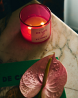 A lit ROEN Mexico City Candle by Faire in a glass holder sits on a marble surface next to a heart-shaped pink flower and a partially visible green book with the text "VIE DE C." The candle's label reads "ROSA DE LAS FLORES," evoking the fragrant blooms one might find in La Condesa. The scene is warmly lit.