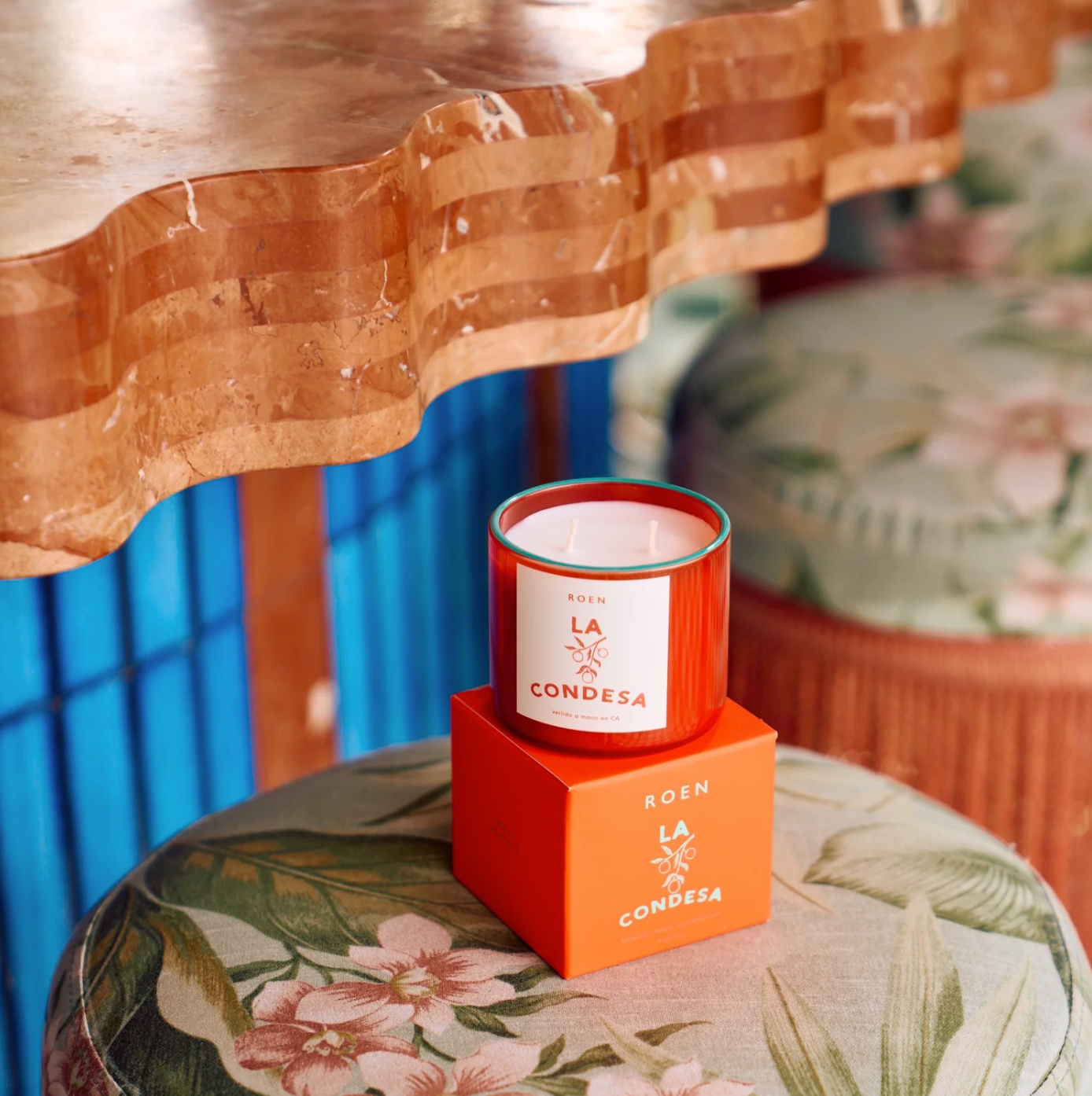 A vibrant orange candle labeled "ROEN Mexico City Candles" by Faire rests on a matching orange box atop a floral-patterned cushion. The backdrop showcases a wavy-edged wooden tabletop with a warm finish and striking blue vertical paneling, evoking the quaint charm of Casa Polanco.