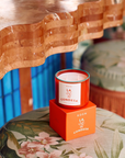 A vibrant orange candle labeled "ROEN Mexico City Candles" by Faire rests on a matching orange box atop a floral-patterned cushion. The backdrop showcases a wavy-edged wooden tabletop with a warm finish and striking blue vertical paneling, evoking the quaint charm of Casa Polanco.