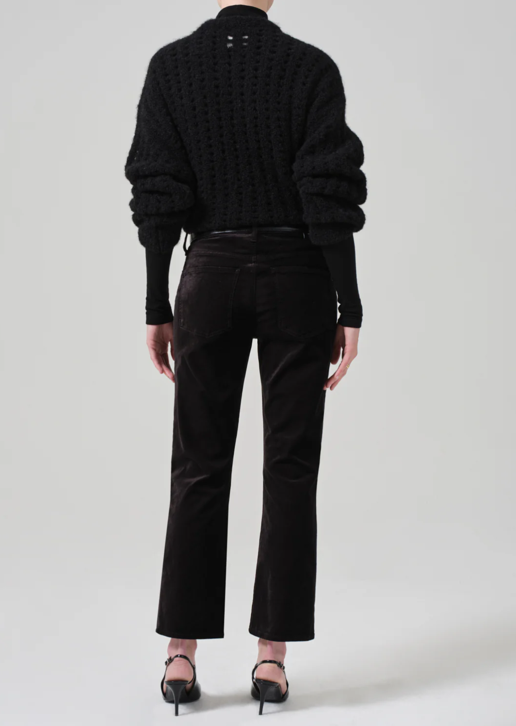 A person is facing away from the camera, showcasing their outfit. They are wearing a chunky black knitted sweater with voluminous sleeves and a mock neck, mid-rise Isola Cropped Trouser in Velvet by Citizens Of Humanity/AGOLDE, and black heeled sandals with ankle straps. The background is plain light gray.