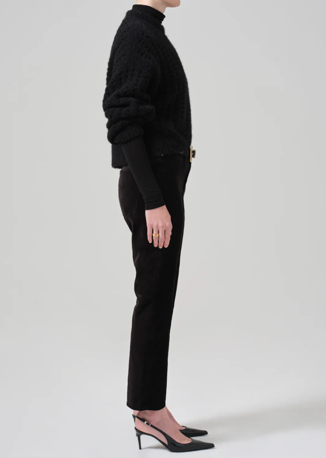 A person standing in a side profile view wearing a black textured sweater, black turtleneck underneath, mid-rise Isola Cropped Trousers in Velvet by Citizens Of Humanity/AGOLDE, and black pointed-toe heels with an ankle strap. The background is plain and light gray.