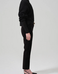 A person standing in a side profile view wearing a black textured sweater, black turtleneck underneath, mid-rise Isola Cropped Trousers in Velvet by Citizens Of Humanity/AGOLDE, and black pointed-toe heels with an ankle strap. The background is plain and light gray.