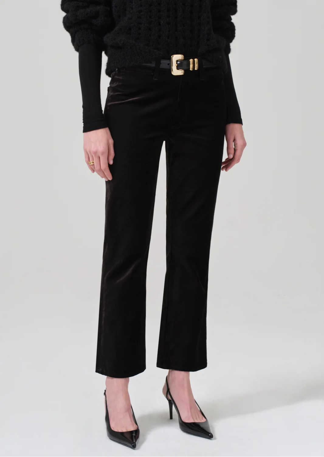 A person stands against a plain background wearing the Isola Cropped Trouser in Velvet by Citizens Of Humanity/AGOLDE, paired with a black pullover and black heeled shoes. They accessorize with a wide belt featuring a large gold buckle and wear a simple gold ring on their left hand.