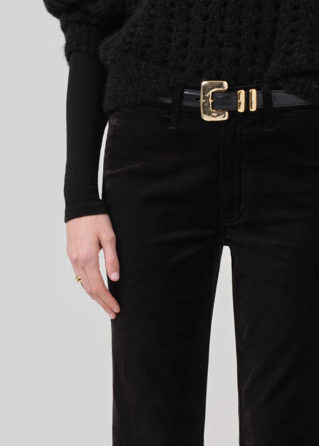 A close-up image of a person wearing a black textured sweater and the Isola Cropped Trouser in Velvet by Citizens Of Humanity/AGOLDE, paired with a gold-buckled black belt. They have their left hand resting beside their hip, adorned with a simple gold ring on their ring finger. The bootcut flare of the velvet trousers adds an elegant touch to the outfit.