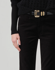 A close-up image of a person wearing a black textured sweater and the Isola Cropped Trouser in Velvet by Citizens Of Humanity/AGOLDE, paired with a gold-buckled black belt. They have their left hand resting beside their hip, adorned with a simple gold ring on their ring finger. The bootcut flare of the velvet trousers adds an elegant touch to the outfit.