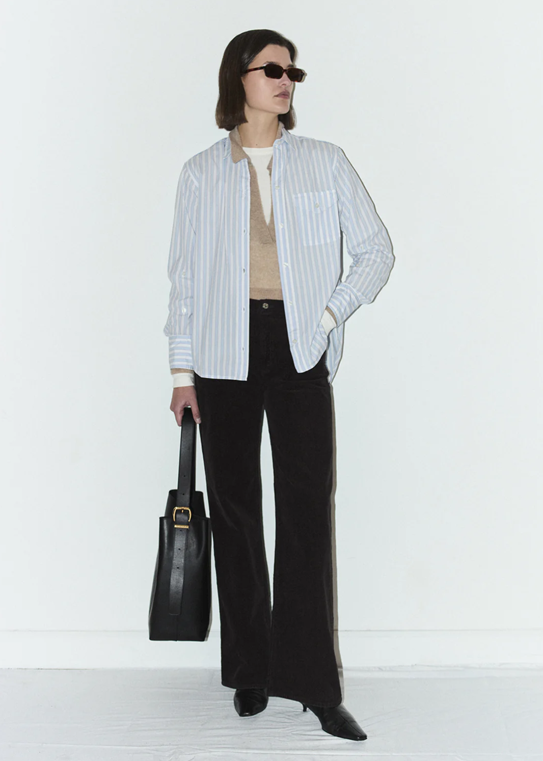 A person with dark hair wearing sunglasses, a striped button-up shirt, and the Annina 30 Zip Fly In Corduroy by Citizens Of Humanity/AGOLDE stands against a plain background. They hold a black tote bag in one hand and have a neutral expression.