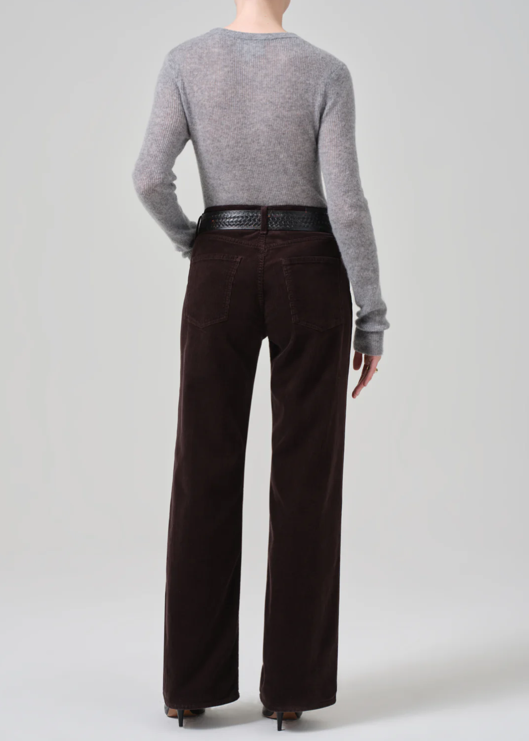 A person with short hair is seen from behind, wearing a light gray long-sleeve sweater tucked into the Citizens Of Humanity/AGOLDE Annina 30 Zip Fly In Corduroy high-waist wide-leg trousers, cinched with a black textured belt. The background is plain light gray.