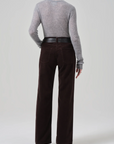 A person with short hair is seen from behind, wearing a light gray long-sleeve sweater tucked into the Citizens Of Humanity/AGOLDE Annina 30 Zip Fly In Corduroy high-waist wide-leg trousers, cinched with a black textured belt. The background is plain light gray.