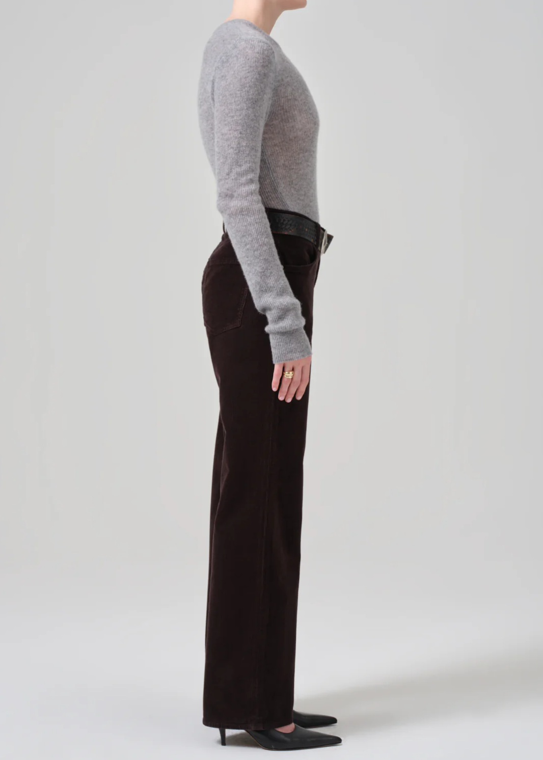 A person stands in profile against a plain background, wearing a long-sleeve grey sweater tucked into high-waist Annina 30 Zip Fly In Corduroy pants by Citizens Of Humanity/AGOLDE. They have on black pointed-toe heels and a black belt. Only the lower part of their face is visible.