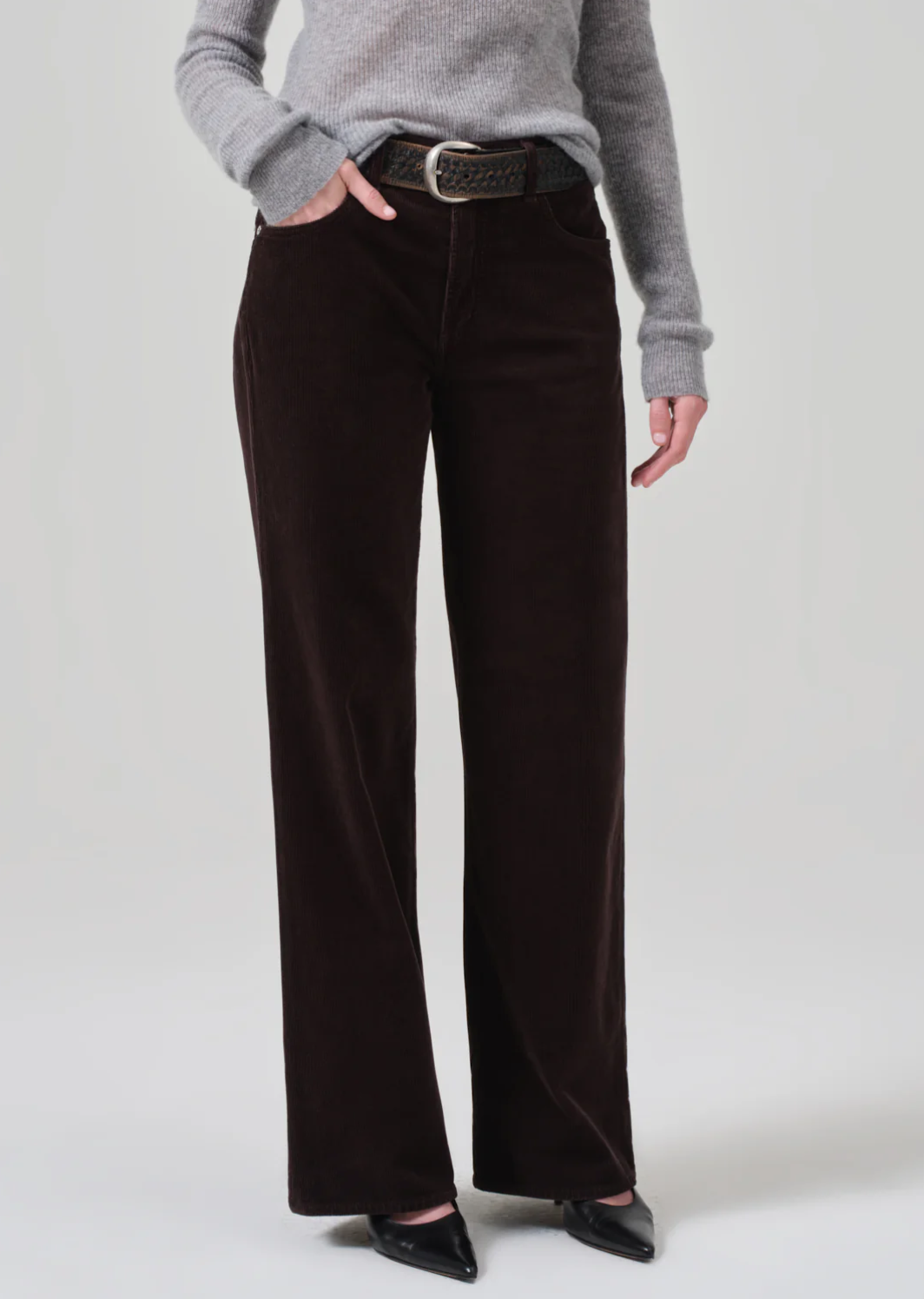 A person stands wearing a gray ribbed sweater, the Annina 30 Zip Fly In Corduroy pants by Citizens Of Humanity/AGOLDE in dark brown with a wide-leg flare, a black woven belt, and black pointed-toe shoes. One hand is in a pocket, and the other rests by their side against a plain background.