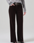 A person stands wearing a gray ribbed sweater, the Annina 30 Zip Fly In Corduroy pants by Citizens Of Humanity/AGOLDE in dark brown with a wide-leg flare, a black woven belt, and black pointed-toe shoes. One hand is in a pocket, and the other rests by their side against a plain background.