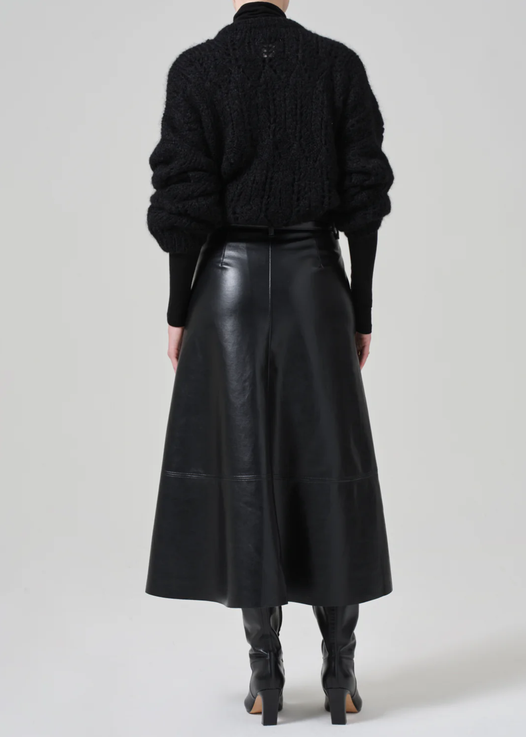 A person is seen from the back, wearing a black textured sweater, the high-rise Cassia Leather Skirt by Citizens Of Humanity/AGOLDE that falls to the knee, and black knee-high boots. The look is monochromatic and chic. The background is plain and light-colored.