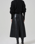 A person is seen from the back, wearing a black textured sweater, the high-rise Cassia Leather Skirt by Citizens Of Humanity/AGOLDE that falls to the knee, and black knee-high boots. The look is monochromatic and chic. The background is plain and light-colored.