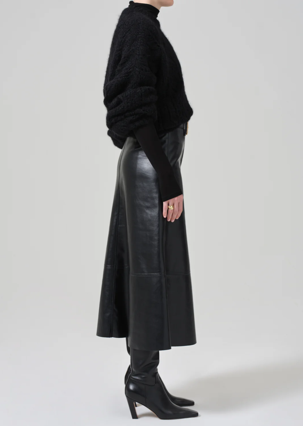 Side view of a person wearing a black textured sweater, the Cassia Leather Skirt by Citizens Of Humanity/AGOLDE, and black boots with pointed toes and heels. The outfit exudes a sleek and stylish aesthetic against a plain, light-toned background.