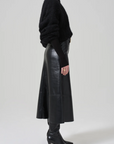 Side view of a person wearing a black textured sweater, the Cassia Leather Skirt by Citizens Of Humanity/AGOLDE, and black boots with pointed toes and heels. The outfit exudes a sleek and stylish aesthetic against a plain, light-toned background.