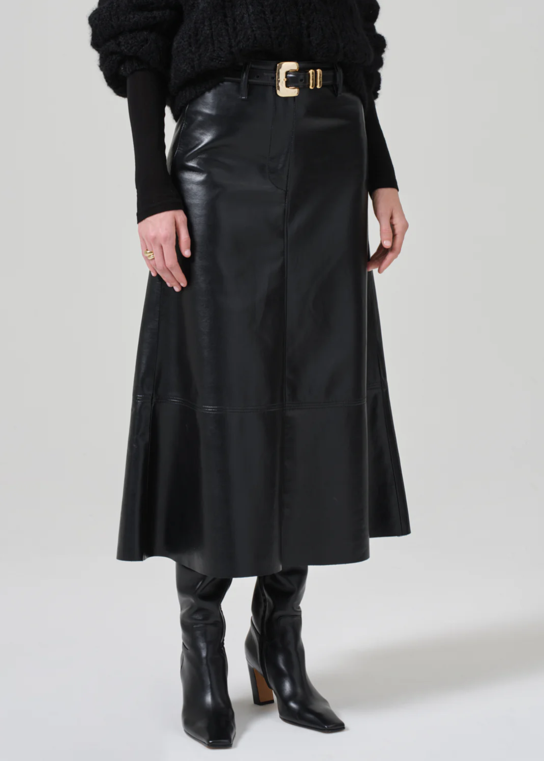 A person is wearing the luxurious **Cassia Leather Skirt** in black from **Citizens Of Humanity/AGOLDE**, featuring a gold-buckled belt, paired with black knee-high boots. They are also wearing a black long-sleeve top with a chunky knit texture. The photo is taken in a well-lit studio against a plain white background.