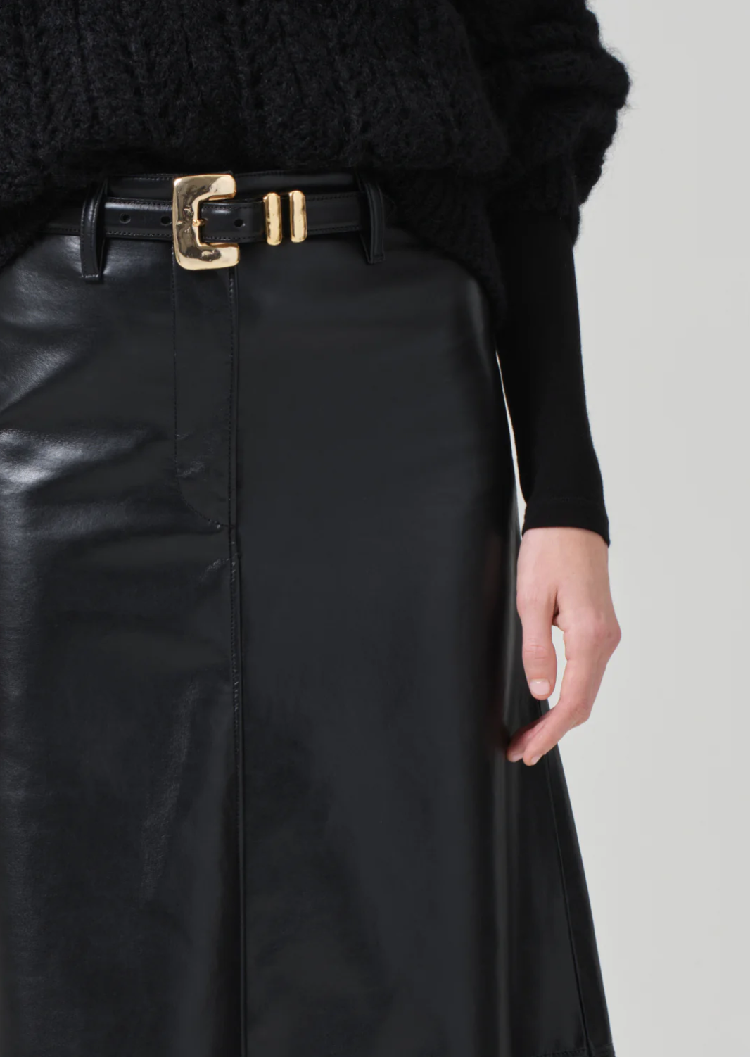 A person wearing the buttery supple Cassia Leather Skirt in black from Citizens Of Humanity/AGOLDE, accessorized with a black belt featuring a gold buckle. The high-rise skirt is paired with a black knitted sweater, and the background is plain light gray.