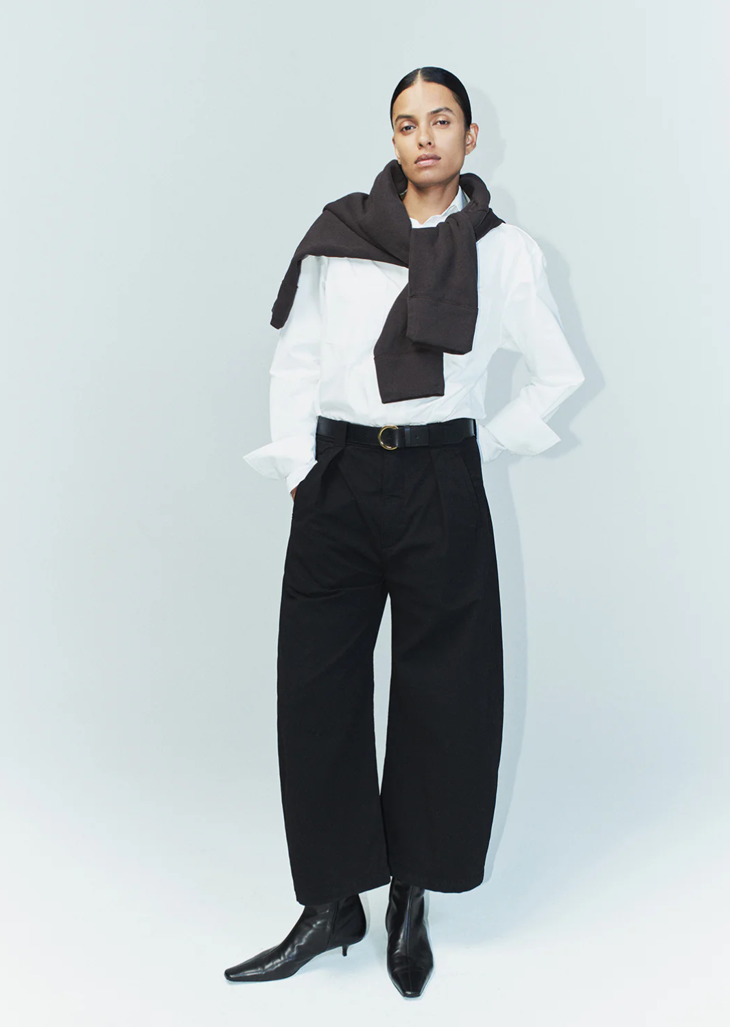 A fashionably dressed person stands against a plain background, sporting a white shirt with a dark sweater draped over their shoulders. They wear the cara cropped pleated trouser in stretch twill by Citizens Of Humanity/AGOLDE and black pointed-toe ankle boots. Their confident stance, with hands in pockets, completes the look.