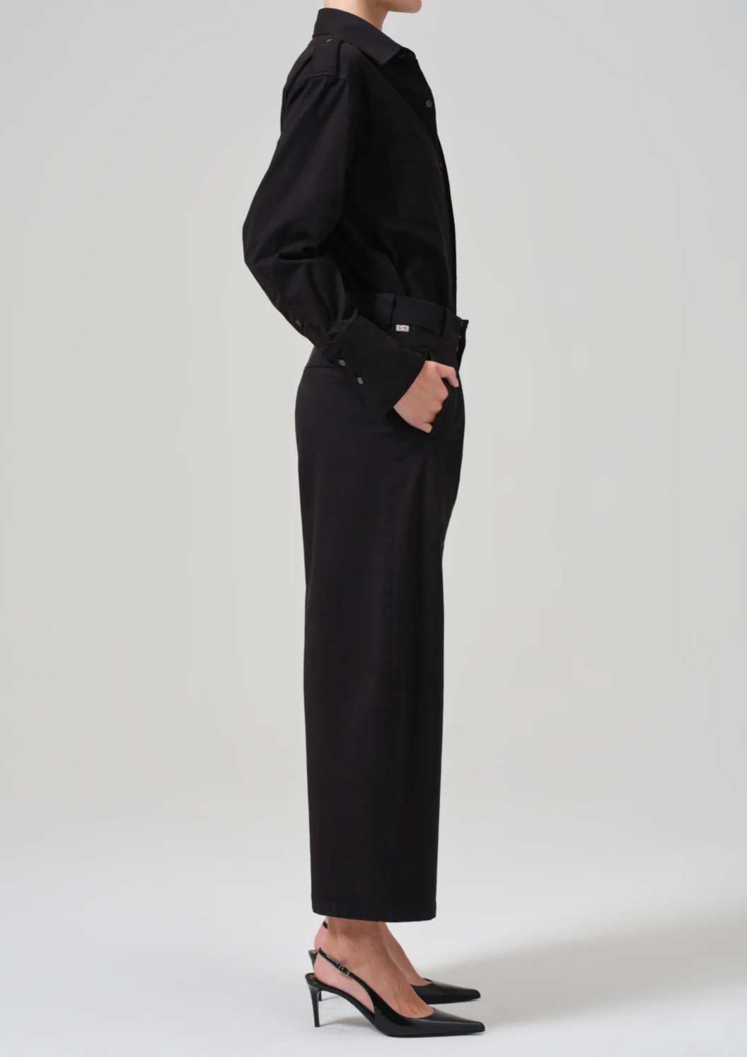 Side profile of a person standing with one hand in their pocket. They are wearing a long-sleeved, button-up black shirt, Cara Cropped Pleated Trouser in Stretch Twill by Citizens Of Humanity/AGOLDE, and black slingback heels with pointed toes. The background is a plain, light gray color.