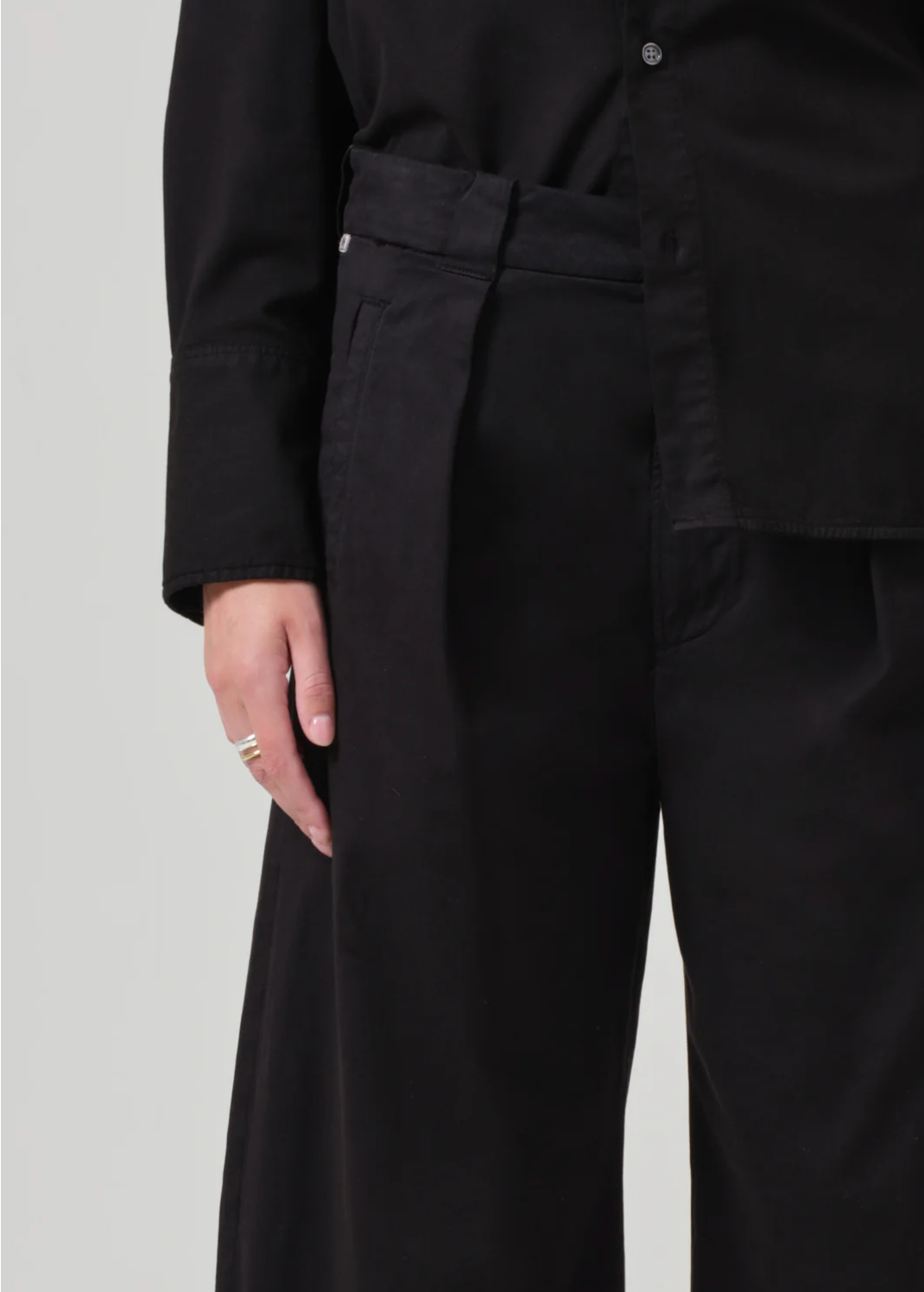 A person dressed in black is shown from the shoulders down. They are wearing a black long-sleeved shirt and black Cara Cropped Pleated Trousers in stretch twill by Citizens Of Humanity/AGOLDE. A silver ring is visible on their left hand, which is resting by their side. The background is plain and light-colored.