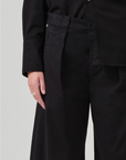 A person dressed in black is shown from the shoulders down. They are wearing a black long-sleeved shirt and black Cara Cropped Pleated Trousers in stretch twill by Citizens Of Humanity/AGOLDE. A silver ring is visible on their left hand, which is resting by their side. The background is plain and light-colored.