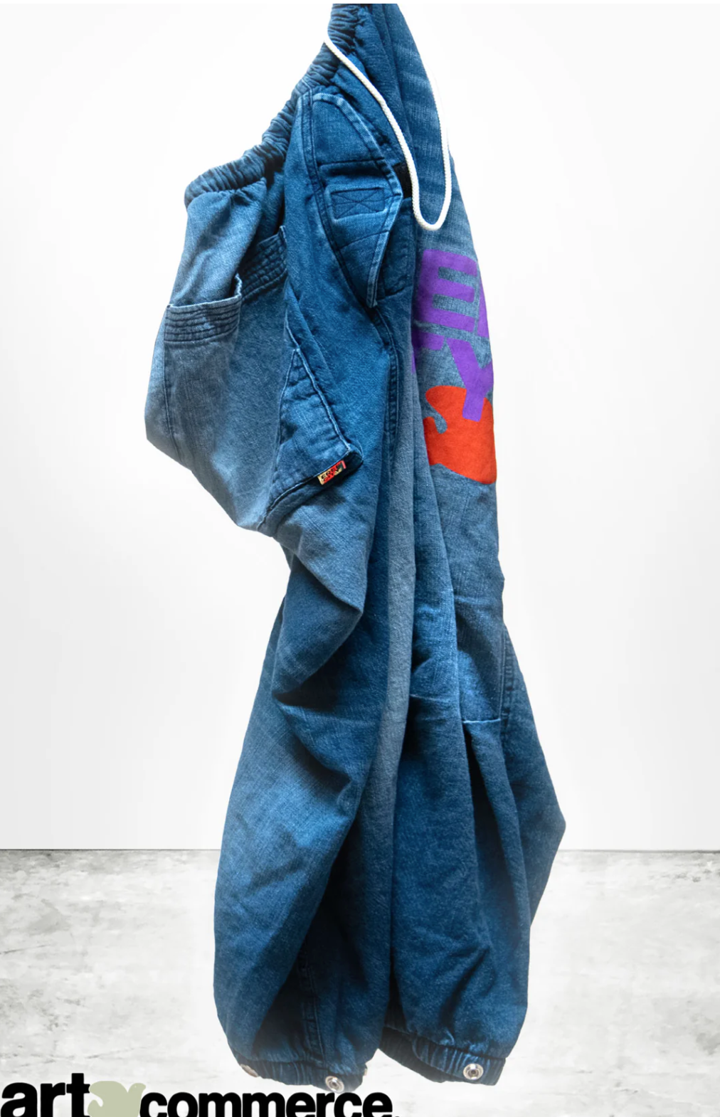 A pair of FLAP/SNAP SHELL FREECITY INDIGO2 jeans, dyed with Japanese indigo, hanging against a white background. The unisex pants feature a colorful patch on one leg, side pockets, and elastic cuffs at the bottom. The tag on the pocket reads "BKE." The "art commerce" logo is partially visible in the bottom left corner. These jeans are from Free City (sparrow, LLC).