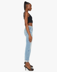 A woman stands in profile against a white background. She has a high ponytail and is dressed in a black tank top, **The Scrapper Cuff Ankle Fray** light blue wash denim high-waisted jeans from **Mother**, and black high-heeled shoes. Her arms are relaxed by her sides.