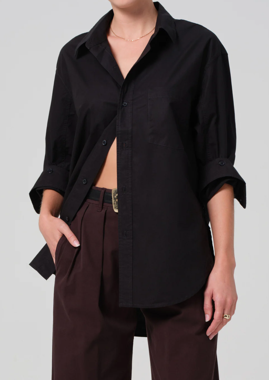 A person wearing a loose-fitting black kayla shirt by Citizens Of Humanity/AGOLDE with the sleeves rolled up to the elbows, and brown pleated pants with one hand in their pocket. The shirt is partially tucked in and they have a thin necklace and a simple ring on one finger.