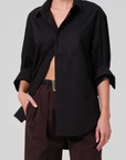 A person wearing a loose-fitting black kayla shirt by Citizens Of Humanity/AGOLDE with the sleeves rolled up to the elbows, and brown pleated pants with one hand in their pocket. The shirt is partially tucked in and they have a thin necklace and a simple ring on one finger.